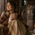 Woman wrapped in Hudson Valley Plaid woven cotton throw blanket holding coffee mug, classic plaid in espresso cider and sage tones with multicolor fringe, warm cabin interior