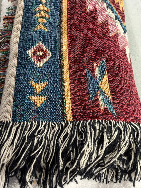 A close-up of a woven textile with fringe, showing a Southwestern geometric pattern in red, blue, beige, and yellow on a white background.