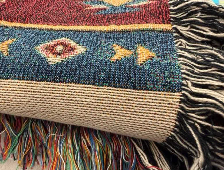 A close-up of a woven textile with fringe, showing a dark blue central band with geometric motifs, a red upper section, a beige lower band, and multicolored tassels.
