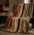 Santa Fe Drift woven cotton throw blanket draped over wooden chair in warm adobe interior, terracotta sage and sand stripe with multicolor fringe