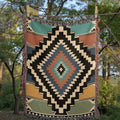 The Sonora Southwestern Cotton Woven Throw Blanket hanging outdoors among trees, showing the full black, terracotta, slate blue, and sage diamond pattern — Trini Skies