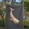 Scrimshaw whale woven cotton throw blanket draped outdoors over a wooden porch railing with green trees in the background, showing the natural texture of the jacquard weave