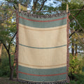 Woman sitting on a beach spread with The Shorewood coastal blue heritage stripe woven cotton throw blanket, reading a book — Trini Skies