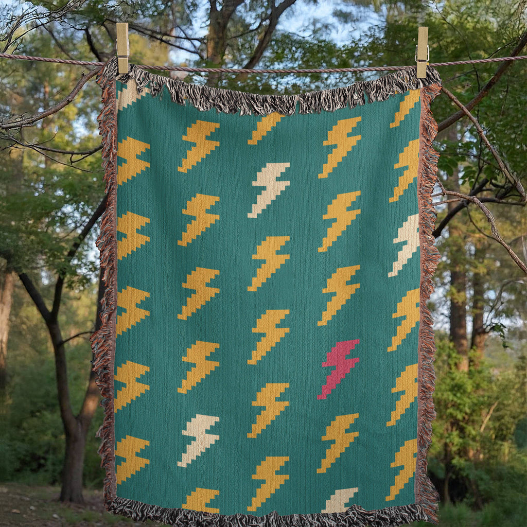Bolt Field woven cotton throw blanket in teal with mustard, cream, and pink lightning bolt scatter pattern, laid flat on grass among trees, full jacquard weave pattern and multicolor fringe visible — Trini Skies