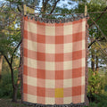 Strawberry gingham woven cotton throw blanket in coral, terracotta, and cream, hanging outdoors on a clothesline among trees, showing the jacquard weave and fringe — Trini Skies