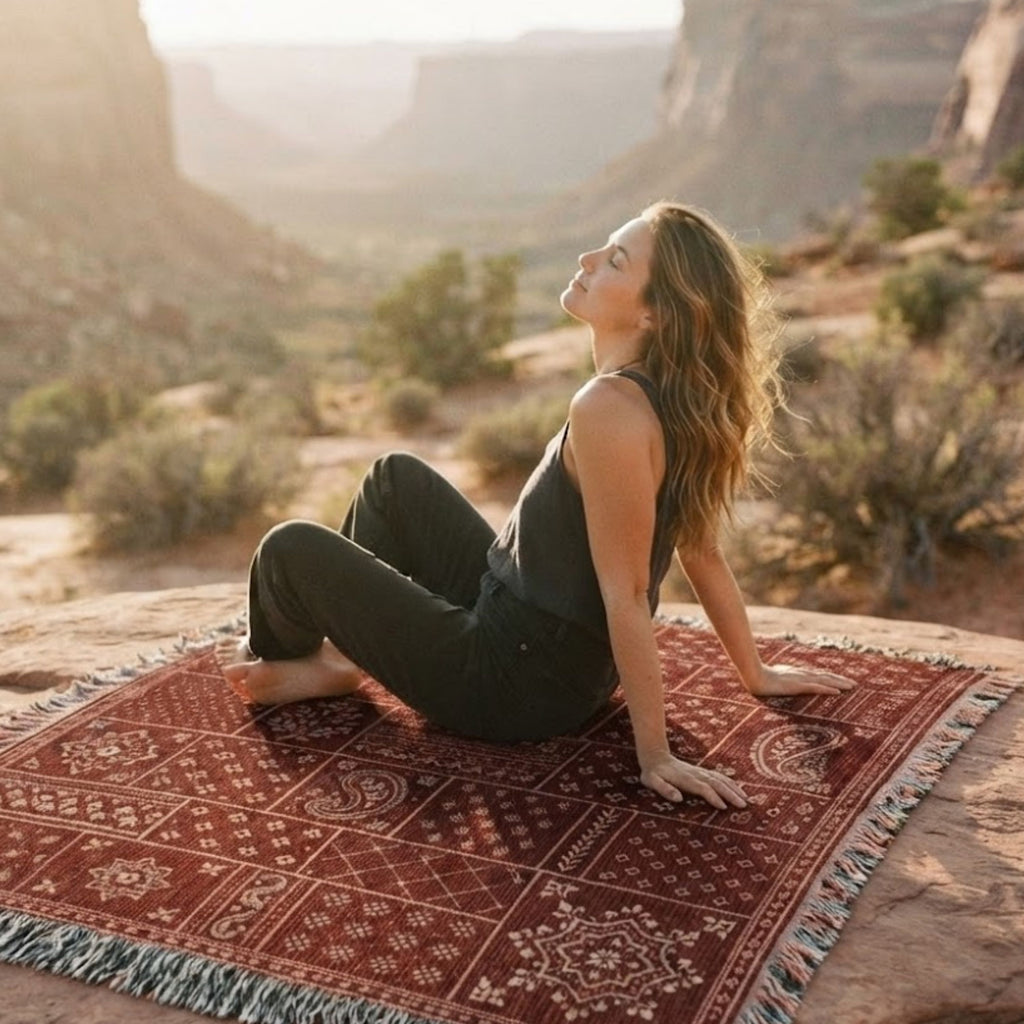 Woman sitting on the Sundown Bandana paisley patchwork woven cotton throw blanket spread across red rock desert terrain — Trini Skies