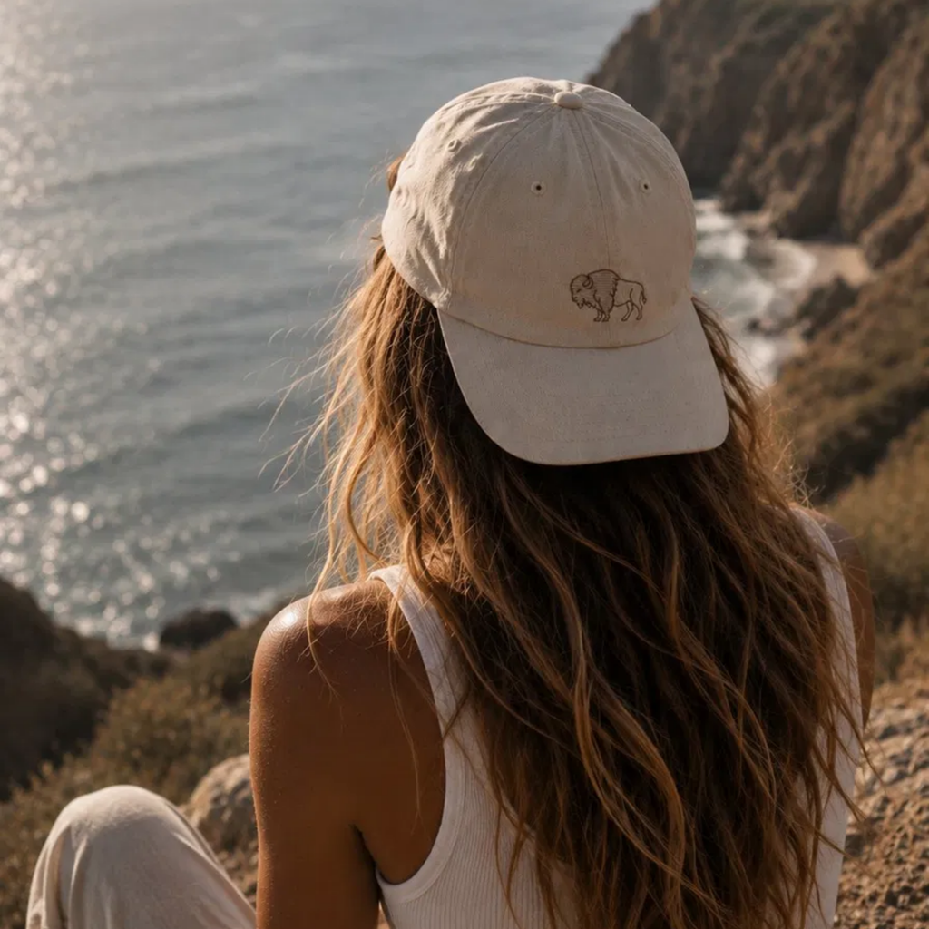 Woman from behind wearing Heritage Bison organic cotton hat, seated on coastal cliff at golden hour overlooking ocean and rocky coastline