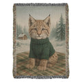 Cozy Lynx Winter Cabin woven cotton throw blanket showing a baby lynx in a forest-green sweater against a snowy cabin scene — Trini Skies