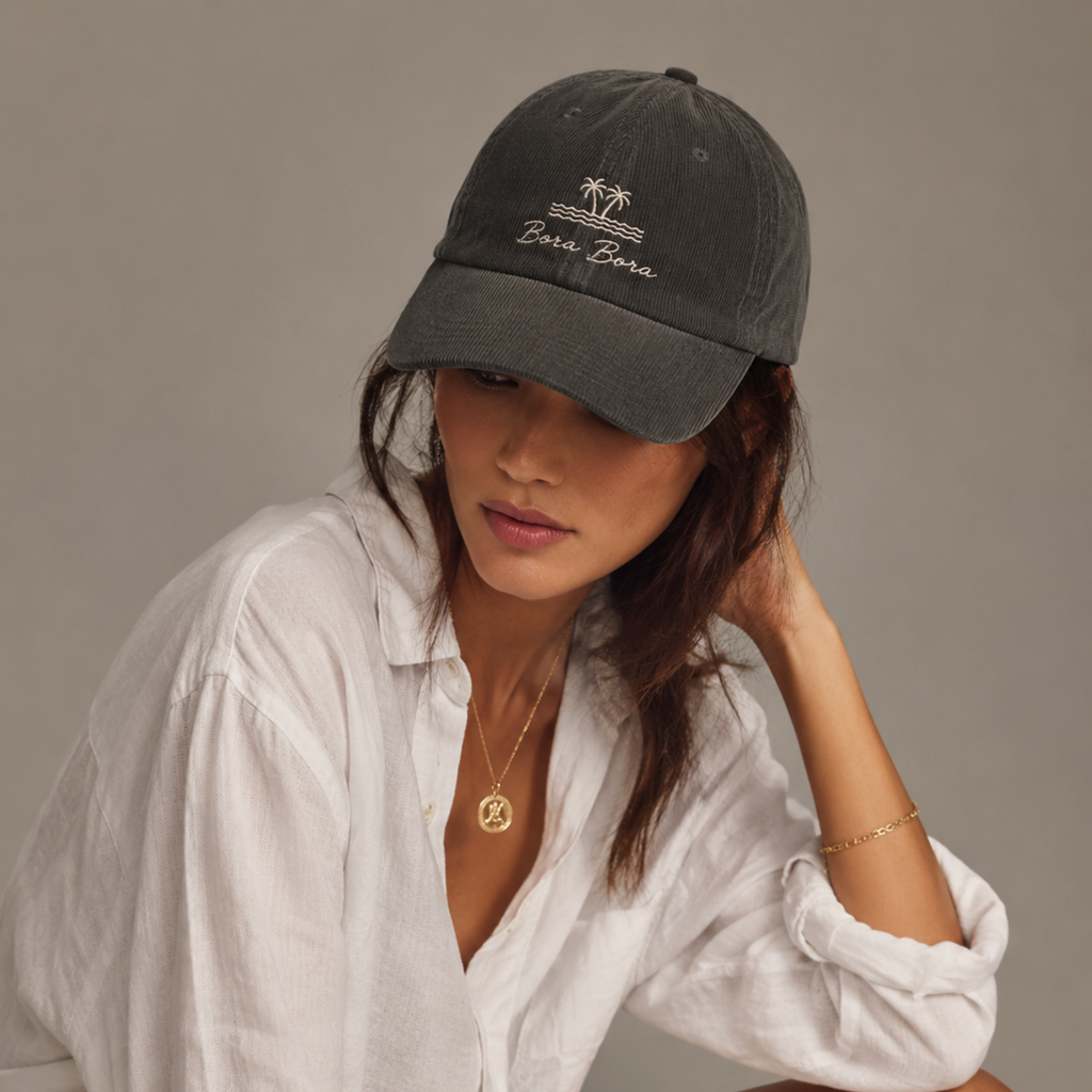 Woman wearing a dark gray cap with 'Bora Bora' text, white shirt, and gold jewelry on a neutral background