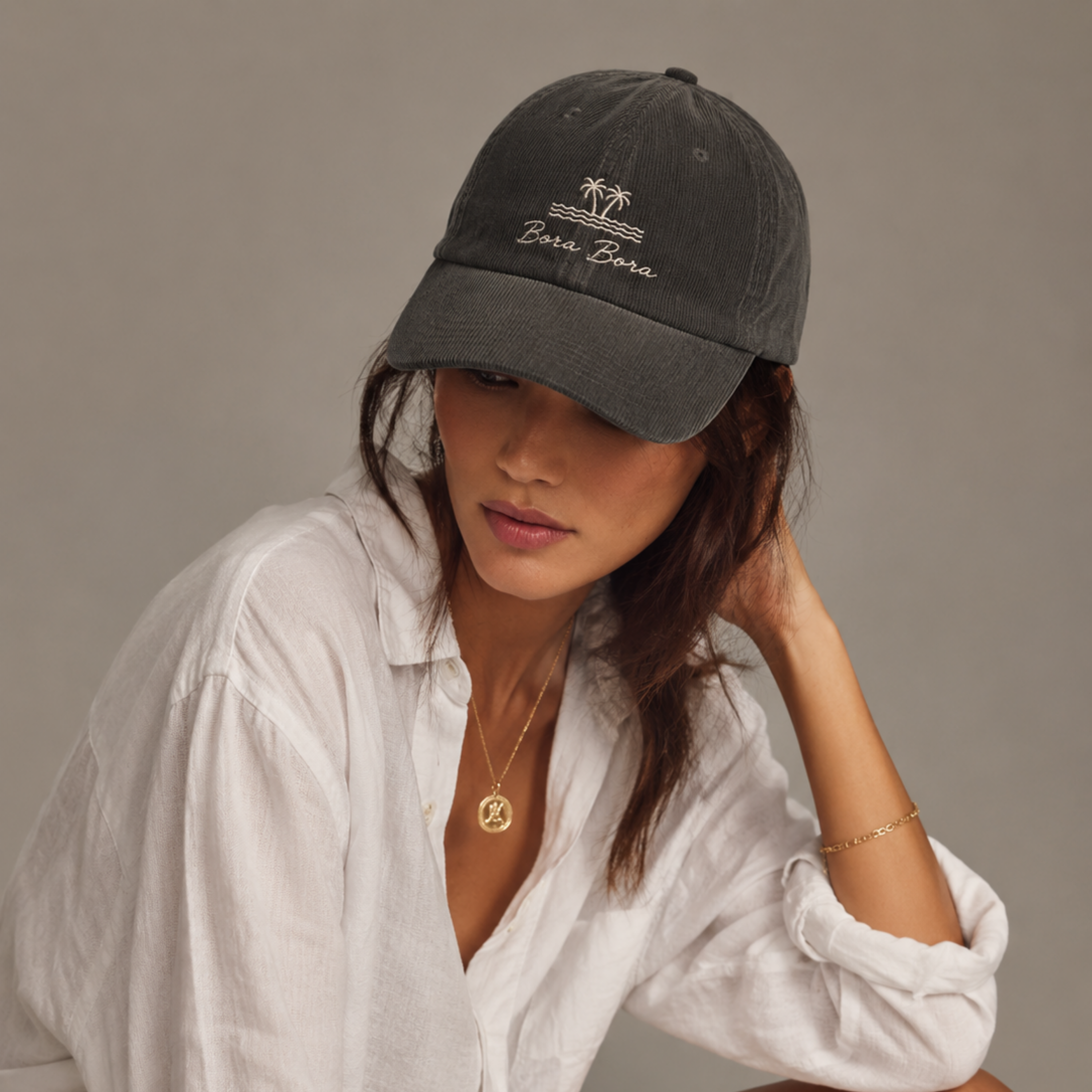Woman wearing a dark gray cap with 'Bora Bora' text, white shirt, and gold jewelry on a neutral background