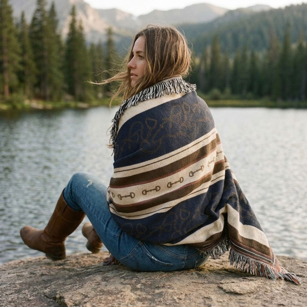 Woman sitting on a rock by a lake wrapped in the Stirrup Row equestrian woven cotton throw blanket, showing the navy and cream stripe pattern and multicolor fringe — Trini Skies