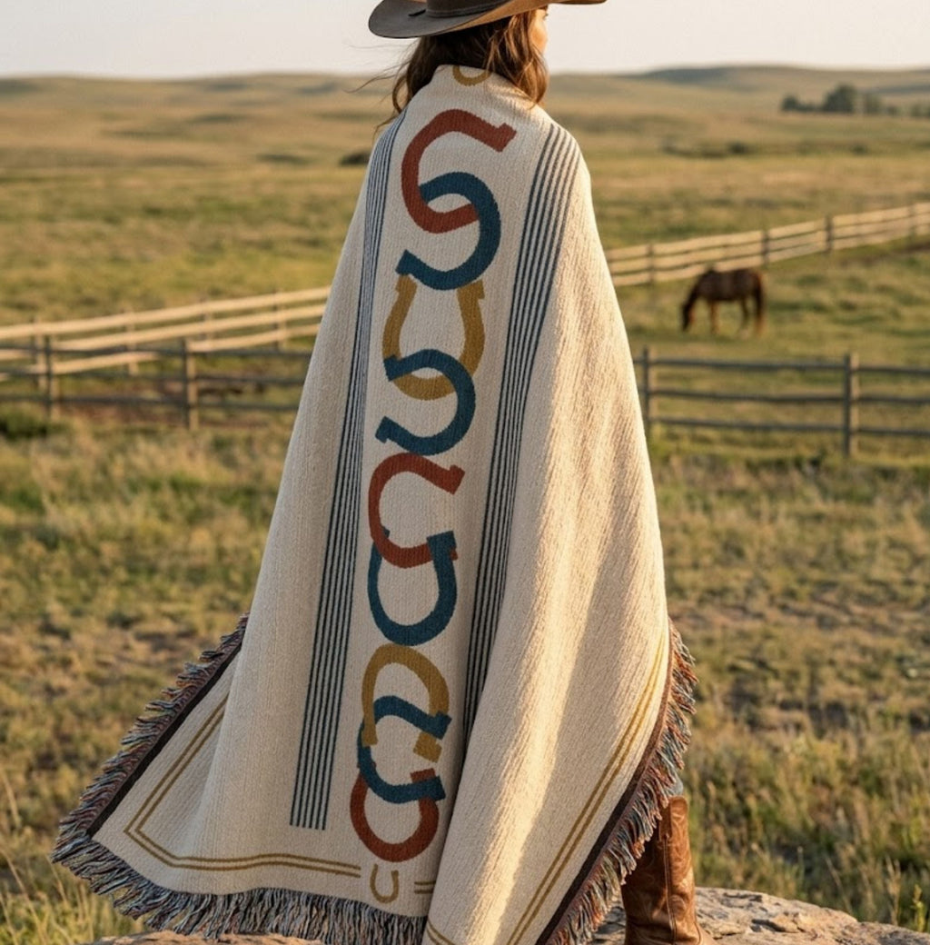 Woman wrapped in the Horseshoe Row woven cotton throw blanket standing in a ranch field with a horse nearby, showing the horseshoe motif pattern in red, rust, and denim blue — Trini Skies