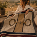 Woman wrapped in Equestrian Estate woven cotton throw blanket at golden hour on a ranch — heritage snaffle bit motif, hunter green burgundy navy stripe, Trini Skies