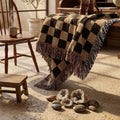 Espresso and oat checkerboard woven throw blanket draped over a wooden stool beside slippers on a cabin porch