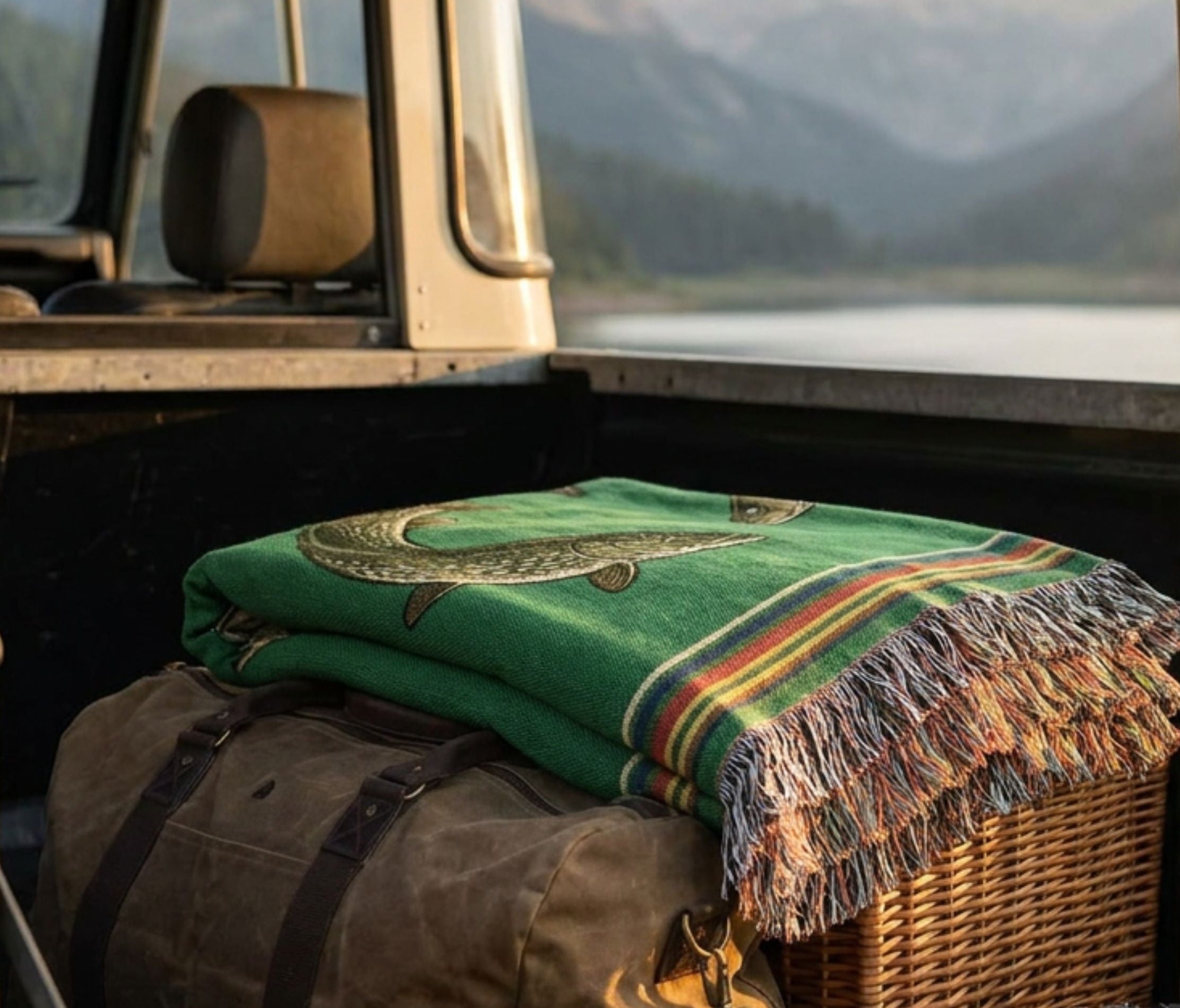 Full view of The Lake Country woven cotton throw blanket, showing the vibrant forest green field with the bass mid-strike, coiled pike, and gliding walleye design, heritage stripe border top and bottom, and multicolor fringe along the sides — Trini Skies