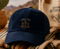 Blue cap with cactus design, beige bag with desert-themed text, and sunglasses on a rustic surface with desert background.