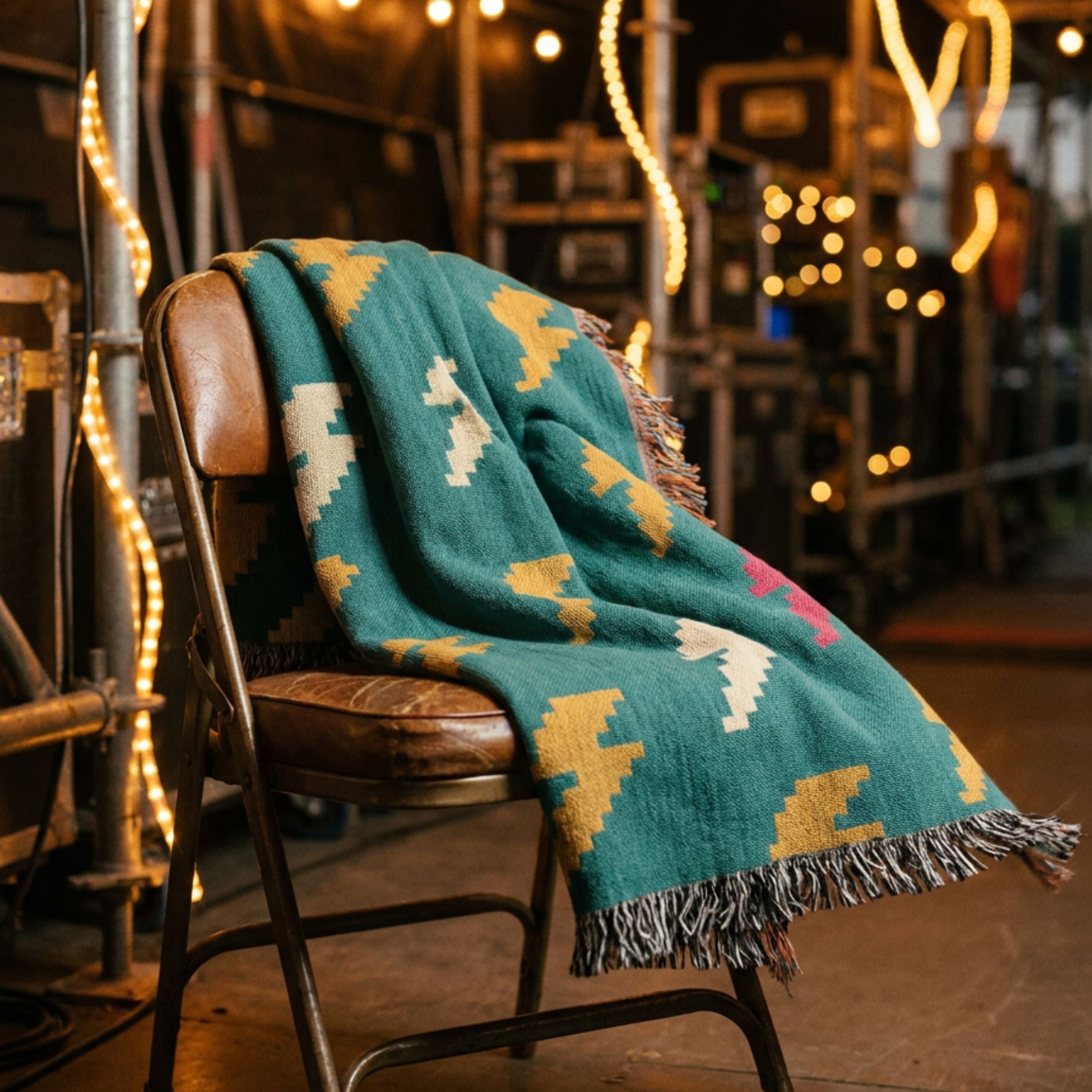 Bolt Field woven cotton throw blanket in teal with mustard, cream, and pink lightning bolt scatter pattern, draped over a wooden chair with string lights glowing in the evening background — Trini Skies