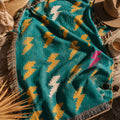 Close-up of the Bolt Field woven cotton throw blanket in teal with mustard, cream, and pink lightning bolts, bunched to show the jacquard weave texture in warm indoor light — Trini Skies