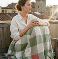 Woman wrapped in the Matcha gingham woven cotton throw blanket in sage green, cream, and coral, sitting on a wooden bench holding a mug, warm golden rooftop light — Trini Skies