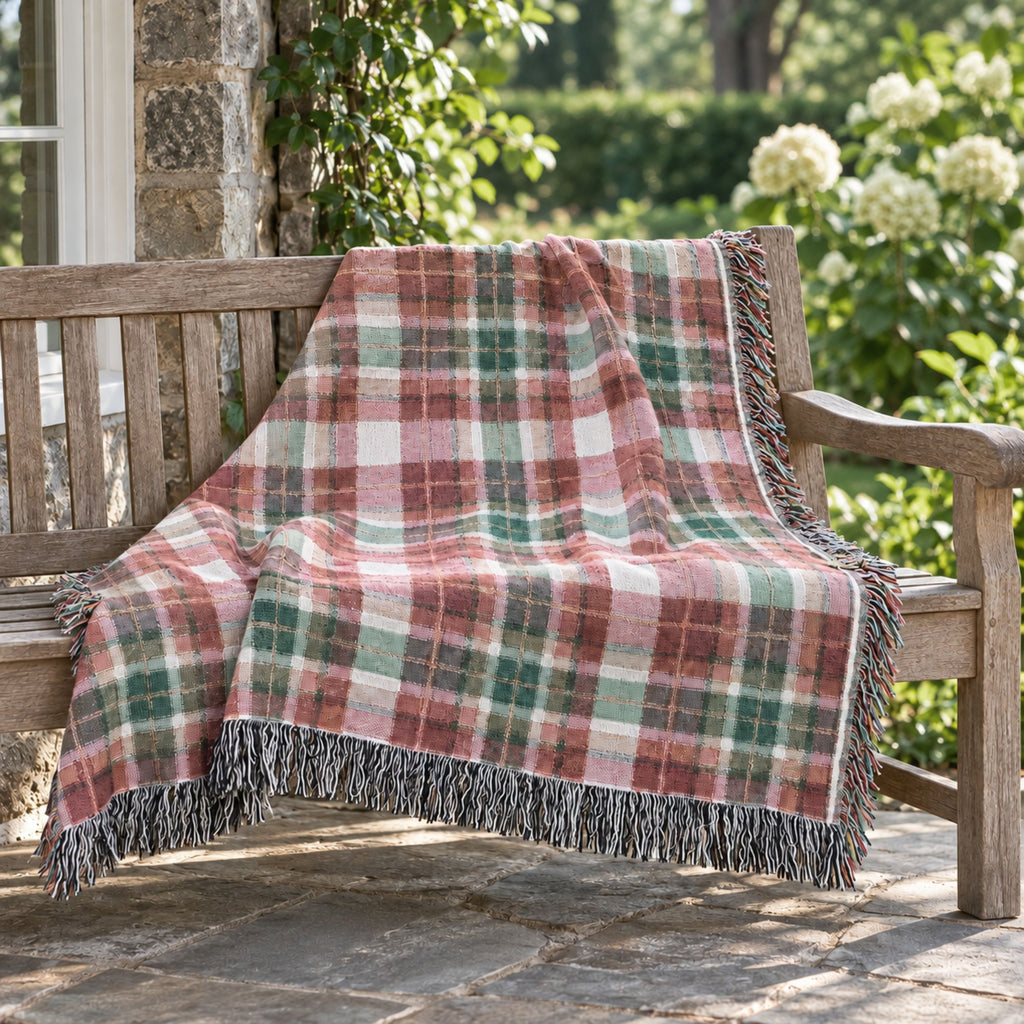 A fringed plaid throw blanket draped over a wooden bench on a stone patio, with green foliage and white flowers in the background.