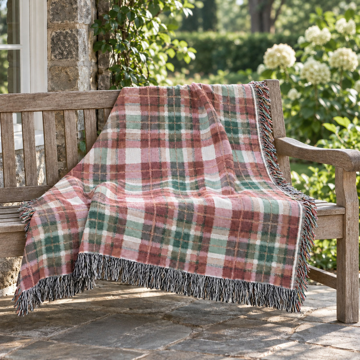 A fringed plaid throw blanket draped over a wooden bench on a stone patio, with green foliage and white flowers in the background.