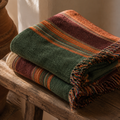 The Ridgeline heritage stripe woven throw blanket folded and stacked on a rustic wooden table, burgundy, forest green, rust, and tan stripe pattern with multicolor fringe — Trini Skies