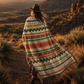 Woman wrapped in the Mesa Serape Southwest woven cotton throw blanket — warm ivory serape stripes and Saltillo diamonds — standing in open desert landscape at golden hour — Trini Skies