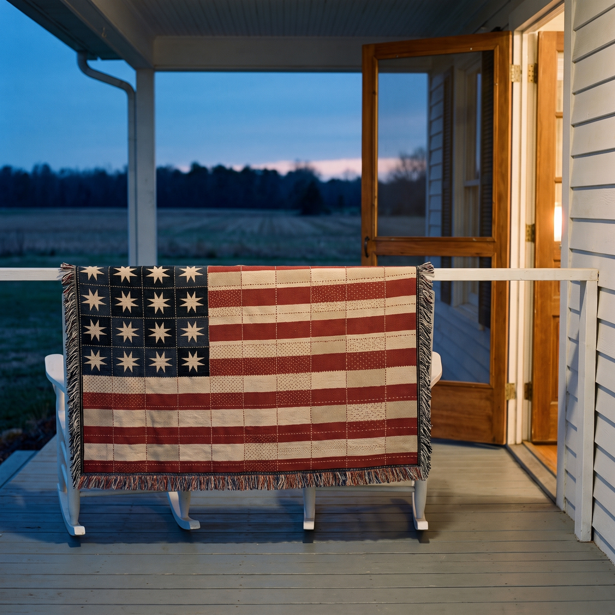 A rustic American flag-themed quilted blanket draped over a white rocking chair on a porch at dusk, with a house interior glowing in the background.