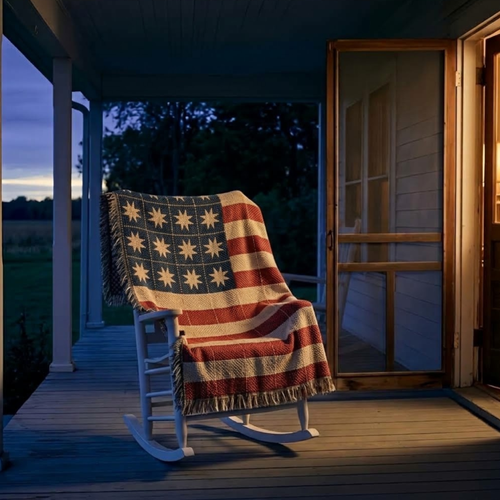 Quilt-style American flag cotton throw draped over a white porch rocker at dusk, lamplight from the open door
