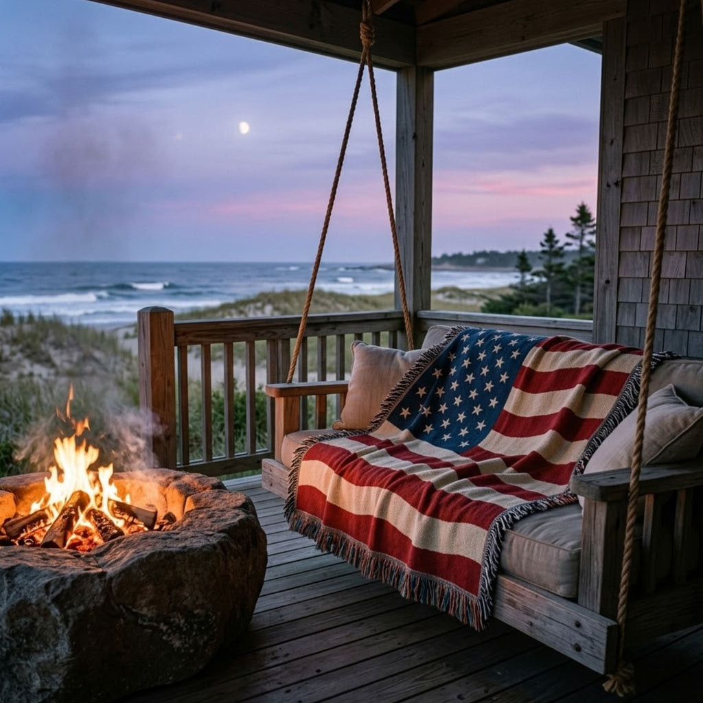 A rustic wooden porch swing draped with an American flag throw blanket, set on a wooden deck with a stone fire pit and ocean view at dusk.