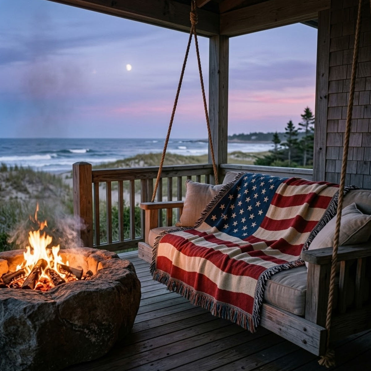 A rustic wooden porch swing draped with an American flag throw blanket, set on a wooden deck with a stone fire pit and ocean view at dusk.
