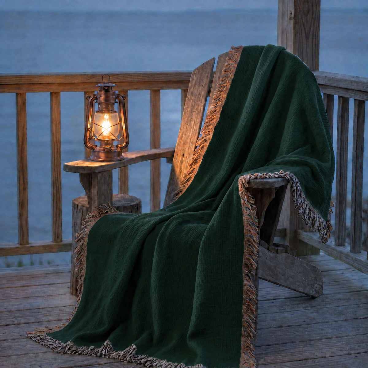 Solid forest green woven cotton throw blanket draped over a wooden chair on a porch at dusk, warm lantern light and multicolor side fringe visible — Trini Skies