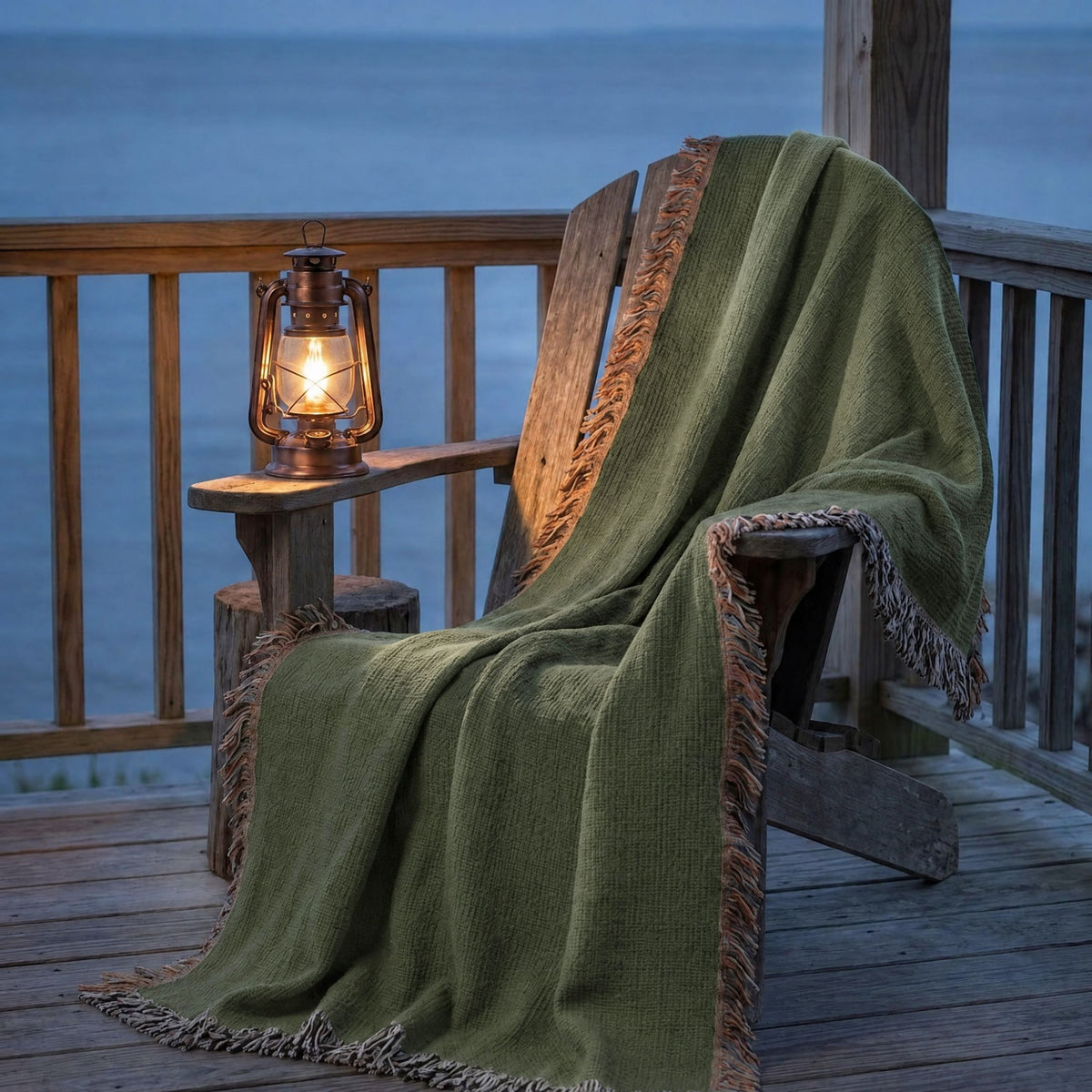 Moss green cotton throw blanket draped over a porch chair beside a glowing lantern above a lake at twilight