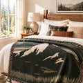 A mountain and forest patterned throw blanket draped over a bed in a sunlit bedroom. The blanket is dark charcoal green with cream and tan woven motifs, and the bed is styled with white linens, plaid and brown accent pillows, a wooden headboard, a bedside lamp, and a window showing trees outside.