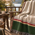 Ivory and forest green colorblock woven cotton throw blanket with multicolor fringe draped over a wooden porch chair overlooking a lake