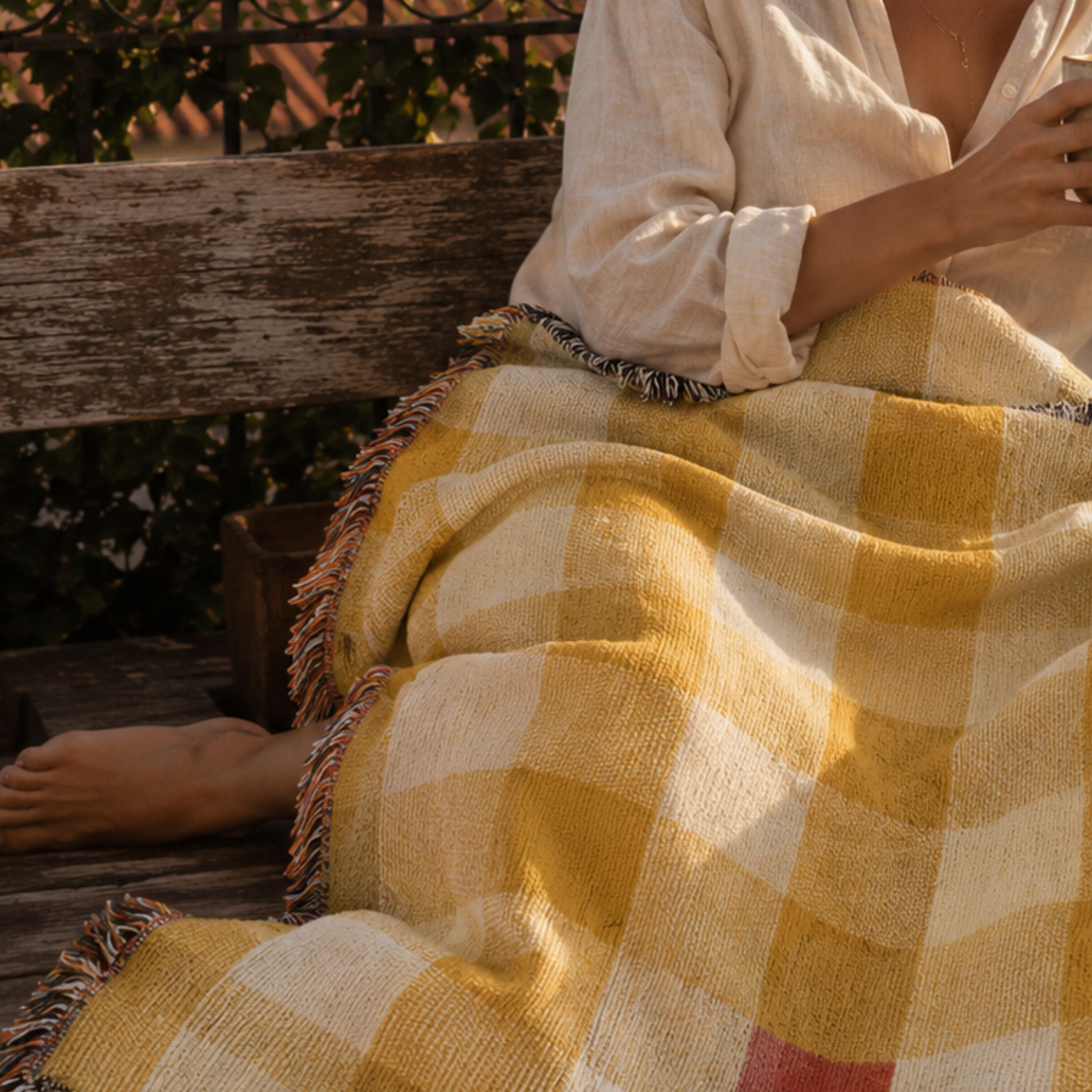 Woman wrapped in the Butter gingham woven cotton throw blanket in mustard gold and cream, holding a coffee mug in warm golden-hour light — Trini Skies